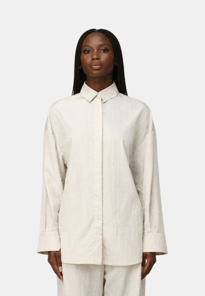 Donna con capelli lunghi intrecciati indossa una camicia ampia color avorio con bottoni e pantaloni abbinati su uno sfondo grigio chiaro uniforme.