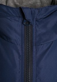 Granatowa kurtka puchowa o strukturalnym wykończeniu, z centralnym czarnym zamkiem błyskawicznym z okrągłym elementem oraz wysokim kołnierzem.