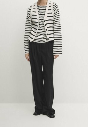 Striped black and white long-sleeve shirt under a cream buttoned cardigan, paired with loose black trousers and flat black shoes.