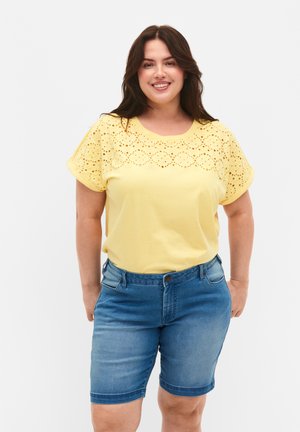 T-shirt di cotone gialla con design a occhietti sulle spalle, abbinata a pantaloni corti in denim blu, con una vestibilità standard e tasche.