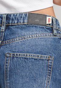 Jeans de denim azul con un diseño de pierna ancha, con costuras naranjas y una etiqueta de cuero negro en la cinturilla que detalla la marca.