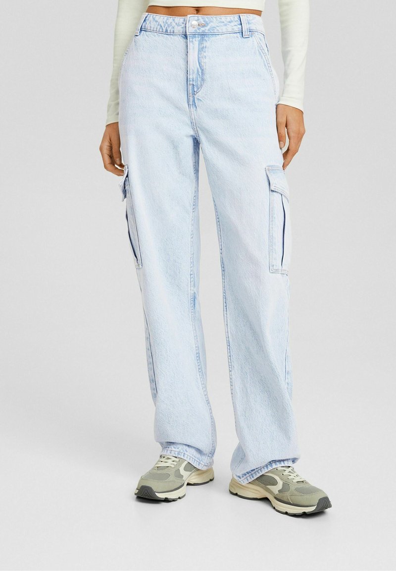 Pantalon cargo en denim bleu clair à jambes larges avec poches latérales, porté avec des baskets beige et grises et un haut court à manches longues.