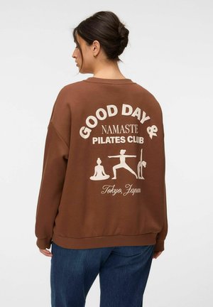 Femme aux cheveux foncés en chignon porte un sweat-shirt marron avec "Good Day Namaste Pilates Club Tokyo, Japan" et des illustrations de postures de yoga dans le dos.