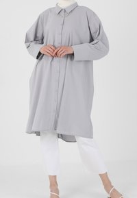 Robe chemise gris clair à boutons avec col, manches longues et poches latérales. Un pantalon blanc court et des sandales simples à lanières complètent le look.