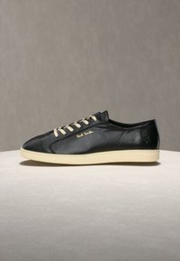 Baskets basses en cuir noir avec lacets blancs, semelle crème et logo doré "Paul Smith" sur le côté, présentées sur une surface claire.
