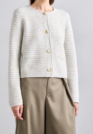 Frau trägt eine strukturierte cremefarbene Strickjacke mit goldenen Knöpfen über beigen, hoch taillierten, weit geschnittenen Hosen.