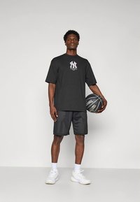 Fekete rövid ujjú póló grafikus logóval, fekete hálós rövidnadrággal párosítva. A modell kosárlabdát tart, fehér tornacipőt visel.