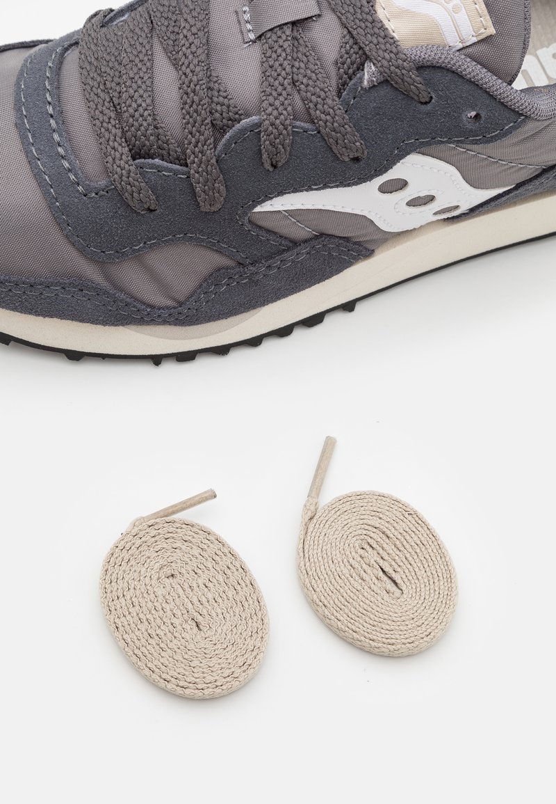 Sneaker grigio realizzato in materiali in nylon e camoscio, con logo bianco, lacci piatti e due lacci beige arrotolati accanto.