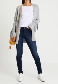 Szary otwarty kardigan z długimi rękawami, biała koszulka, ciemnoniebieskie skinny jeans i białe sneakersy. Żółta torba na ramię z detalem w postaci frędzli.