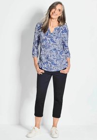 Blaue Bluse mit Paisleymuster, V-Ausschnitt und Dreiviertelärmeln, kombiniert mit schwarzen, knöchellangen Hosen und weißen Sneakers.