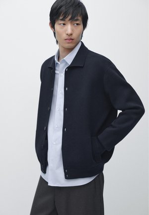 Jeune homme portant un cardigan bleu marine boutonné par-dessus une chemise blanche et un pantalon foncé, debout avec une main dans la poche, sur fond blanc.