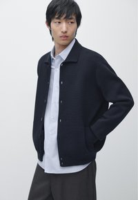 Junger Mann trägt eine marineblaue, geknöpfte Strickjacke über einem weißen Hemd und dunkler Hose, steht mit einer Hand in der Tasche vor weißem Hintergrund.