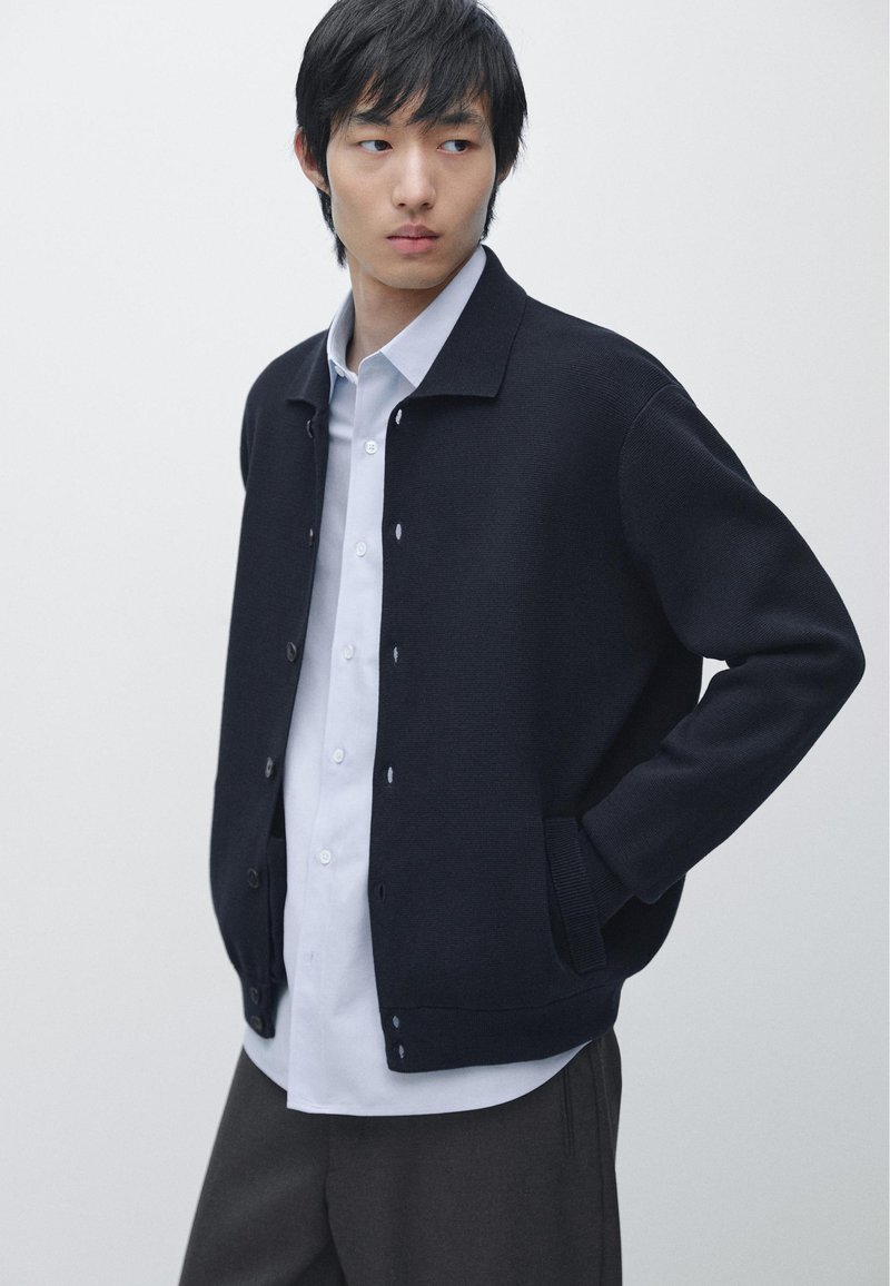 Junger Mann trägt eine marineblaue, geknöpfte Strickjacke über einem weißen Hemd und dunkler Hose, steht mit einer Hand in der Tasche vor weißem Hintergrund.