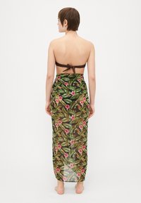 Blomstrete omslagskjørt med gjennomsiktig design som har grønne blader og rosa blomster, brukt over en brun bandeau-top. Bakside.