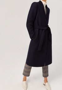 Cappotto avvolgente in lana navy con cintura, maniche lunghe e revers larghi, abbinato a pantaloni cropped a quadri e mocassini bianchi.