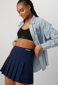 Mornarsko plisirana suknja s adidas logotipom na struku, uparena s crnim sportskim grudnjakom i svijetloplavom prugastom majicom.
