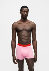 Moški model s temno poltjo nosi roza boksarice z rdečim pasom, na katerem piše "HAPPY", na enobarvnem svetlo sivem ozadju.