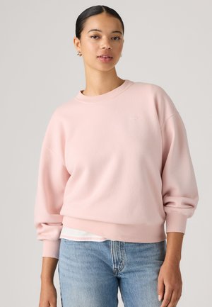Ung kvinde med mørkt hår samlet bagpå iført en lyserød sweatshirt og blå jeans, stående mod en ensfarvet baggrund.