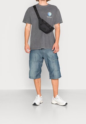 Šedé oversized tričko, denimové cargo šortky, černá crossbody taška a bílé tenisky. Pohodlný outfit s různými texturami a barvami.