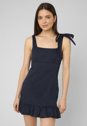 Femme portant une robe bleu marine sans manches avec un décolleté carré, un ourlet à volants et un nœud noué sur l'épaule gauche, posant devant un fond uni.