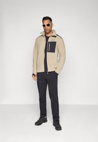 Beige fleece-jakke med en marineblå brystlomme og svarte detaljer, parret med svarte bukser og svarte sko. Modellen har på seg solbriller.