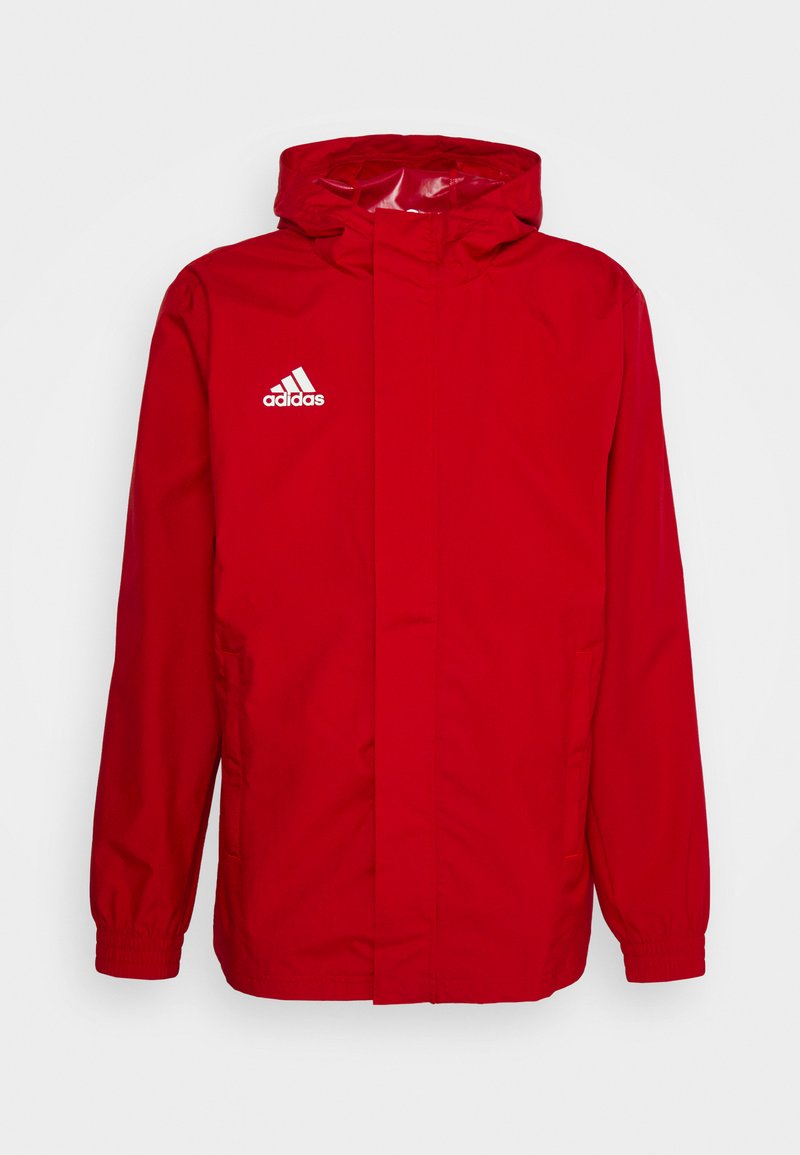 Giacca impermeabile rossa con cappuccio, logo Adidas bianco sul lato sinistro del petto, polsi elasticizzati e tasche laterali. Texture liscia, design casual.