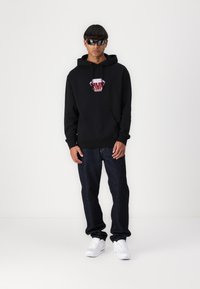 Sort hættetrøje med et rødt logo, parret med mørke denim jeans og hvide sneakers. Modellen bærer solbriller.