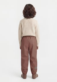 Copil cu păr creț stând cu fața spre spate, purtând un pulover bej tricotat, pantaloni maro cu talie elastică și pantofi maro pe fundal alb.