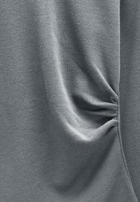 Szary sweter z miękkiej dzianiny, o subtelnej fakturze i marszczonym wykończeniu po bokach, tworzący luźny fason.