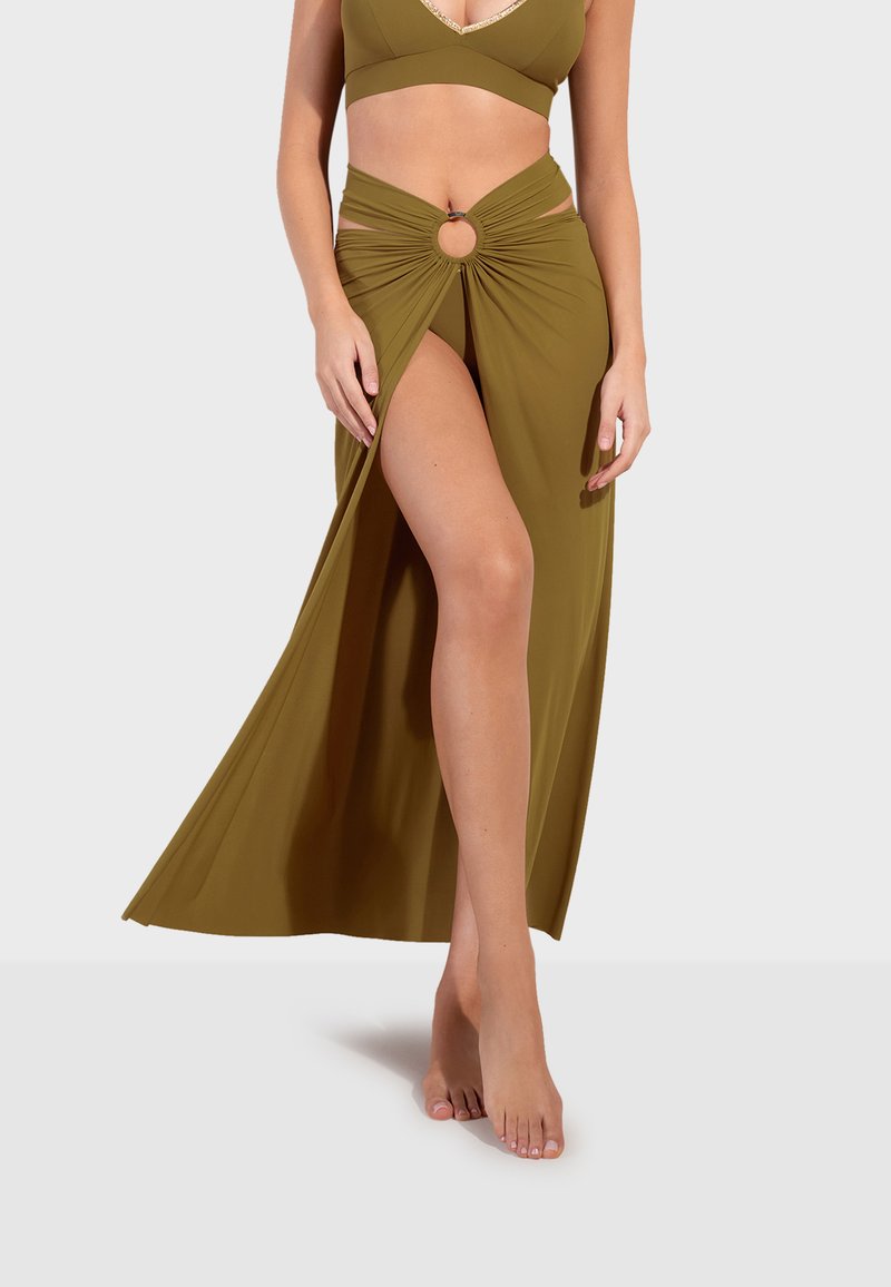 Femme portant une tenue deux pièces vert olive avec un détail d'anneau à la jupe fendue haute, debout pieds nus devant un fond clair.
