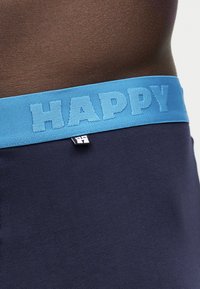 Približan posnetek temne kože v pasu, oblečene mornarsko modro spodnje perilo z svetlo modrim pasom z vtisnjenim napisom "HAPPY" in majhno črno-belo etiketo.