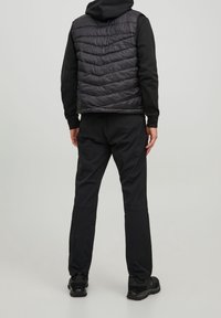 Gilet nero imbottito con un motivo a chevron testurizzato, indossato sopra un felpa nera. Pantaloni e scarpe nere completano l'outfit, con polsini aderenti alle caviglie.