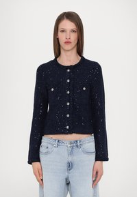 Marineblå cropped jakke med teksturert stoff, frontlommer og sølvknapper. Har rund halslinning og subtile metalliske detaljer.