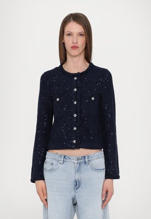 Giacca accorciata blu navy in tessuto strutturato, con tasche frontali e bottoni argentati. Presenta un collo rotondo e sottili accenti metallici.
