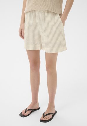 Beine tragen beigefarbene gestreifte Shorts mit elastischem Bund und schwarze Flip-Flops, Hände teilweise in den Taschen, stehen auf weißem Hintergrund.