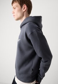 Dark gray hoodie made of soft fabric, features a large hood, ribbed cuffs, and a subtle embroidered logo on the front.