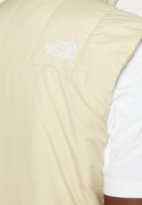 Gilet beige senza maniche con logo bianco "The North Face" sul petto, indossato sopra una camicia bianca a maniche corte.