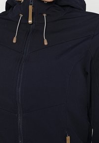 Veste softshell bleu marine dotée d'une fermeture éclair frontale, d'une capuche réglable avec cordons, et d'un tissu texturé avec des accents contrastants couleur beige.