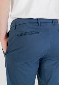 Pantaloni aderenti di un blu scuro, con una texture liscia e una leggera lucentezza. Due tasche posteriori con pattine, frontale piatto e passanti per cintura.