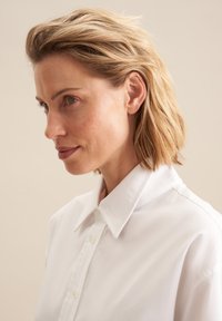 Witte blouse met een gladde textuur en knopen, aangevuld met een ontspannen ontwerp en subtiele glans, gedragen door een persoon met kort haar.