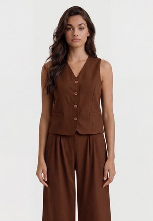 Donna con capelli lunghi e scuri che indossa un gilet marrone senza maniche con bottoni e pantaloni a gamba larga abbinati, in piedi davanti a uno sfondo semplice.