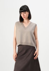 Woman with short dark hair wearing a beige sleeveless V-neck sweater vest and a dark brown skirt standing against a white background.