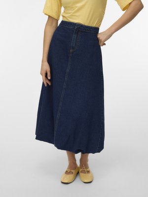 Personne portant un haut jaune, une jupe midi en jean bleu foncé avec ourlet bouffant, et des ballerines jaunes avec des nœuds, debout sur un fond clair.