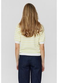 Žena s dlouhými blond vlasy, oblečená v bílé a žluté pruhované krátké tričko a tmavě modré kalhoty, otočená zády proti jednoduchému pozadí.