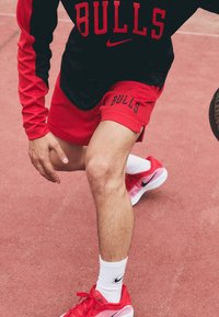Red mesh basketball shorts with "BULLS" lettering, paired with a black long-sleeve top. Hot pink Nike shoes and white socks. Basketball court background.