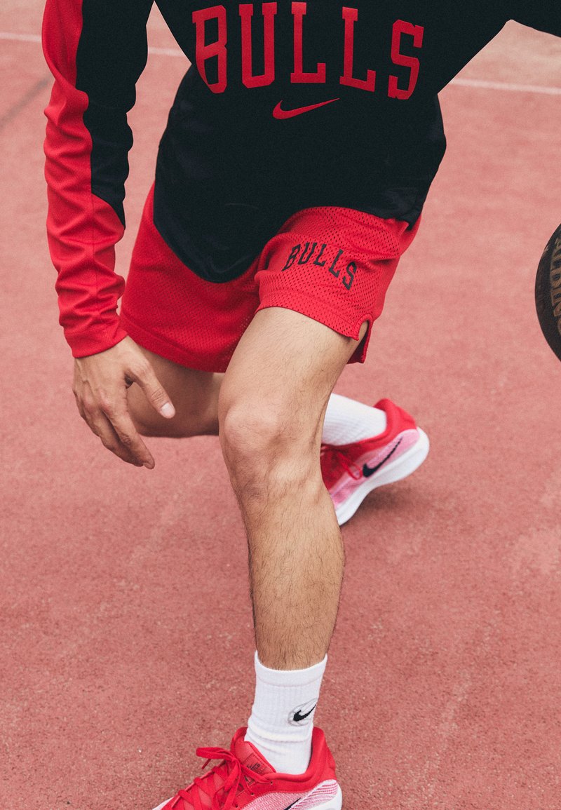Red mesh basketball shorts with "BULLS" lettering, paired with a black long-sleeve top. Hot pink Nike shoes and white socks. Basketball court background.