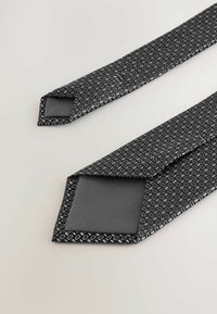 En sort slips med et struktureret mønster i grå og hvid, der har en glat, ensfarvet sort underside og en spids ende.