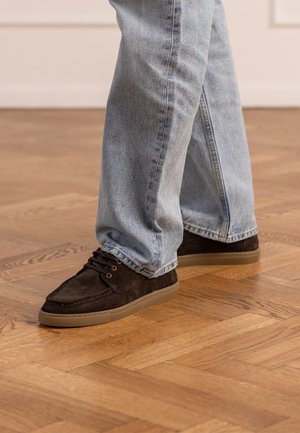 Persona che indossa jeans azzurri chiari e scarpe stringate in pelle scamosciata marrone scuro, in piedi su un pavimento in parquet di legno.