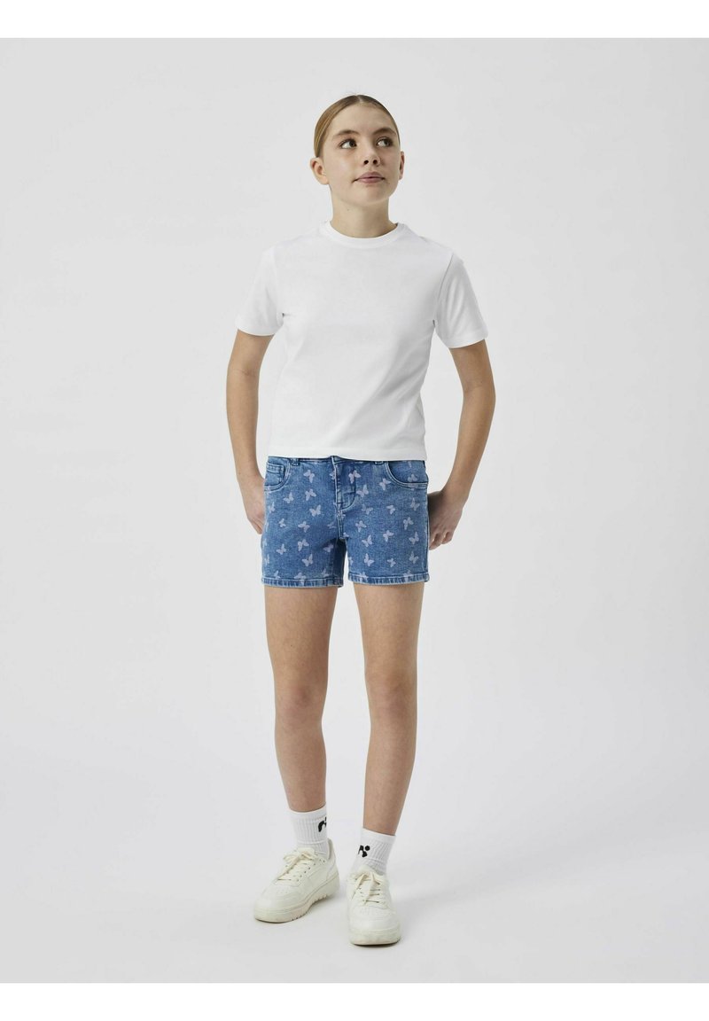 T-shirt blanc à manches courtes associé à un short en denim avec un motif de papillons. Des baskets blanches et des chaussettes mi-mollet complètent le look.