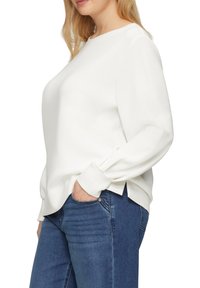 Witte top met lange mouwen en een ronde hals, zachte textuur en zijspleten, gecombineerd met donkerblauwe jeans. Eenvoudig, hedendaags ontwerp.